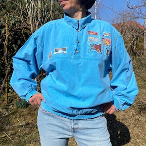Può includere: Una camicia vintage turchese con collo alto e diverse toppe. La camicia ha maniche lunghe con polsini elasticizzati e vita con coulisse. La persona indossa jeans azzurri e occhiali da sole.