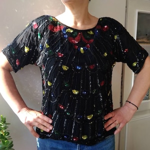 Puede incluir: Una blusa negra de lentejuelas con un diseño floral colorido. La blusa tiene mangas cortas y un escote redondo.