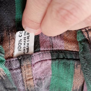 May include: Close-up of a fabric label sewn into a garment. The label is white with black text that reads "100% CC", "Hand Made in", and "Wash Cold".
