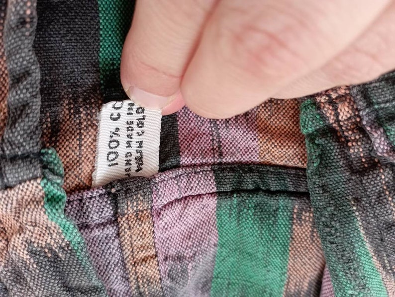 May include: Close-up of a fabric label sewn into a garment. The label is white with black text that reads "100% CC", "Hand Made in", and "Wash Cold".