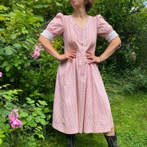 Puede incluir: Un vestido rosa con estampado floral con mangas cortas y cuello en V. El vestido tiene un corpiño ajustado y una falda acampanada, con pequeños botones en la parte delantera. Las mangas están adornadas con encaje blanco. La persona lleva botas de vaquero negras.