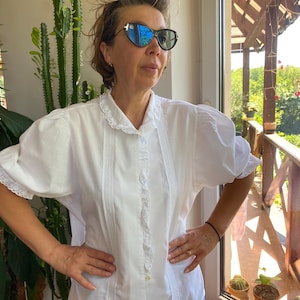 Puede incluir: Blusa blanca con mangas cortas abullonadas y cuello con encaje. La blusa presenta detalles verticales de encaje en la parte delantera y está abotonada. Las mangas y el cuello tienen delicados ribetes de encaje. La persona lleva gafas de sol.