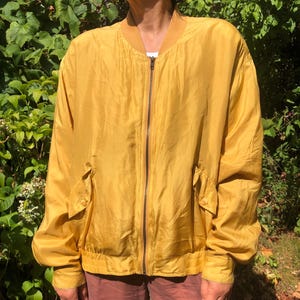 Könnte beinhalten: Eine senfgelbe Bomberjacke mit durchgehendem Frontreißverschluss, geripptem Kragen und gerippten Ärmelbündchen. Die Jacke verfügt über zwei Seitentaschen mit geknöpften Klappen und einen gerippten Saum aus einem leichten, leicht glänzenden Stoff.
