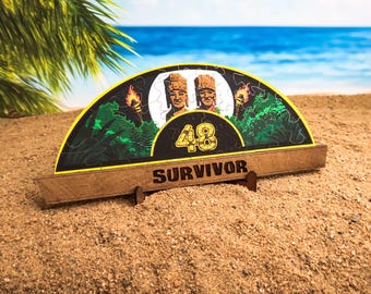 Könnte beinhalten: Ein halbrunder Holzpuzzle-Teil mit schwarzem Hintergrund und einer grünen Dschungelszene. Das Puzzle-Teil zeigt zwei Figuren mit gelben Gesichtern und braunen Haaren, die Zahl 48 und das Wort "SURVIVOR".