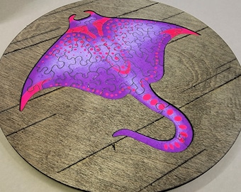 Könnte beinhalten: Ein lila und pinkfarbenes, stingrayförmiges Holzpuzzle auf einem Holzgrund. Das Puzzle ist zusammengesetzt und zeigt den Körper und den Schwanz des Stachelrochens.