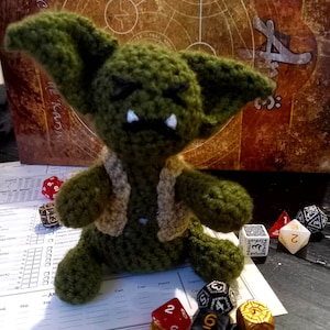Puede incluir: Un juguete de peluche de goblin verde tejido a crochet con un chaleco marrón, sentado en una mesa con dados y un tablero de juego. El tablero de juego tiene el texto "The Karsan".