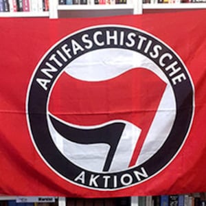 Puede incluir: Una bandera roja con un emblema central. El emblema es un diseño en blanco y negro con las palabras "ANTIFASCHISTISCHE AKTION" en un círculo negro. La bandera se muestra frente a una estantería, con libros visibles.