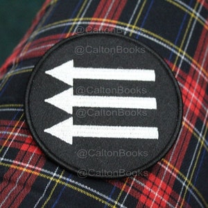 May include: A round black patch with three white arrows pointing to the left. The patch is on a red, black, and yellow plaid fabric. The text "@CaltonBooks" is repeated around the edge of the patch.