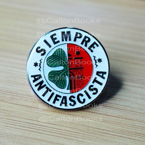 Puede incluir: Un pin blanco redondo con borde negro. El pin presenta un trébol verde, un edificio rojo y el texto "SIEMPRE ANTIFASCISTA" en negro.
