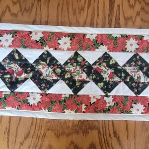 Op de afbeelding: Een witte gestikte tafelrunner met een zwart-wit ruitpatroon en een rode en groene bloemenrand. De runner is ongeveer 40 cm breed en 122 cm lang.