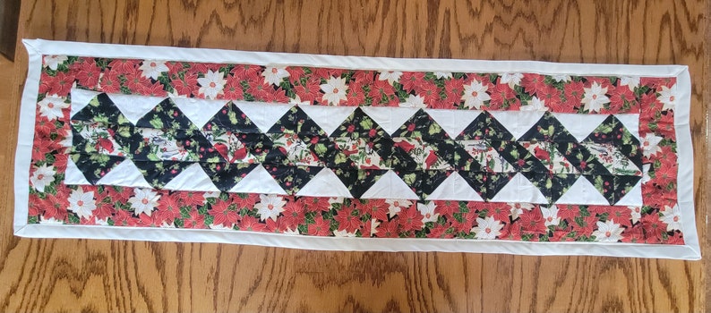 Op de afbeelding: Een witte gestikte tafelrunner met een zwart-wit ruitpatroon en een rode en groene bloemenrand. De runner is ongeveer 40 cm breed en 122 cm lang.