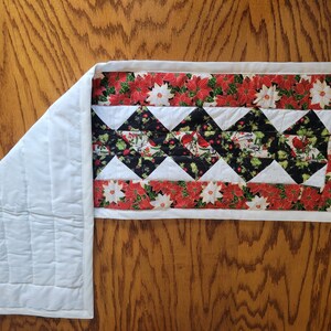 Op de afbeelding: Een witte gestikte tafelrunner met een rode en groene bloemenprint rand en een zwart-wit ruitpatroon in het midden. De runner is ongeveer 40 cm breed en 122 cm lang.