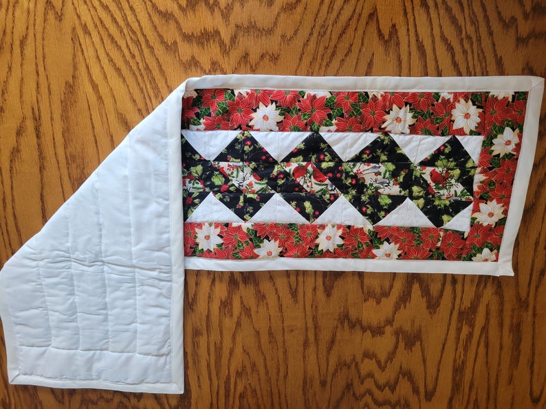 Op de afbeelding: Een witte gestikte tafelrunner met een rode en groene bloemenprint rand en een zwart-wit ruitpatroon in het midden. De runner is ongeveer 40 cm breed en 122 cm lang.