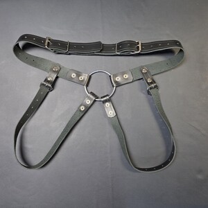 May include: Black leather harness with a large metal ring and adjustable straps. The harness features a belt with two buckles and multiple rivets. The design is symmetrical and appears to be handmade.