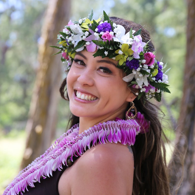 Puede incluir: Una mujer con una corona de flores y un collar de flores. La corona est&aacute; hecha de flores blancas, moradas y verdes. El collar de flores est&aacute; hecho de flores moradas. Ella sonr&iacute;e y mira hacia un lado.