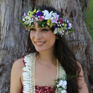 Puede incluir: Una mujer con una corona de flores y un collar de flores blancas, rosas, amarillas y moradas. La corona est&aacute; en su cabeza y el collar alrededor de su cuello. Ella sonr&iacute;e y mira a la c&aacute;mara.