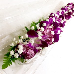 Puede incluir: Un collar de flores morado y blanco hecho con orqu&iacute;deas y aliento de beb&eacute;. El collar est&aacute; dispuesto en una bandeja de pl&aacute;stico transparente.