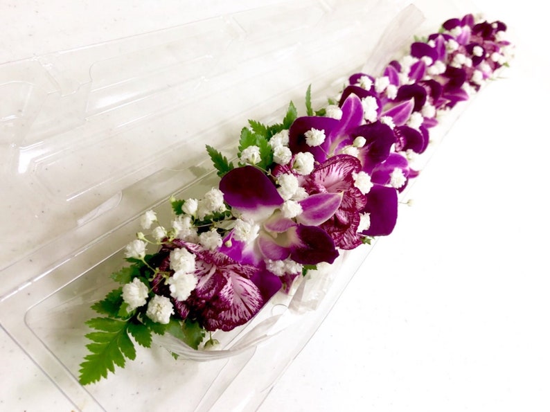 Puede incluir: Un collar de flores morado y blanco hecho con orqu&iacute;deas y aliento de beb&eacute;. El collar est&aacute; dispuesto en una bandeja de pl&aacute;stico transparente.