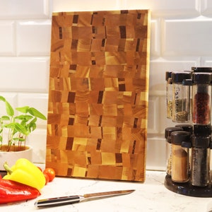 May include: A large wooden cutting board with a unique patchwork design. The board is made of light and dark wood and has a natural finish. It is sitting on a white tiled countertop next to a spice rack.