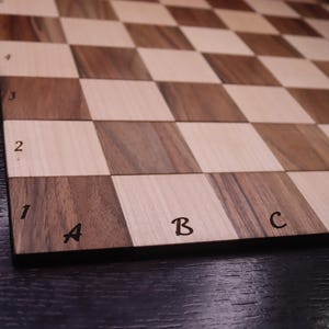 May include: A close-up of a wooden chessboard with alternating light and dark squares. The board has a dark border with the letters A, B, C, and D, and numbers 1, 2, 3, and 4 along the edges. The wood grain is visible.