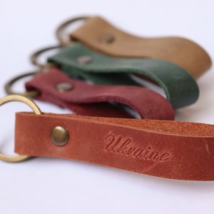 Puede incluir: Cuatro llaveros de cuero con anillos de metal. El llavero de abajo es marrón y tiene la palabra "Ucrania" grabada.