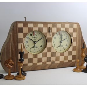 May include: A wooden chess clock with two analog clocks and a checkerboard design. Chess pieces in black and natural wood are arranged in front. The clock face has black hands and Arabic numerals.