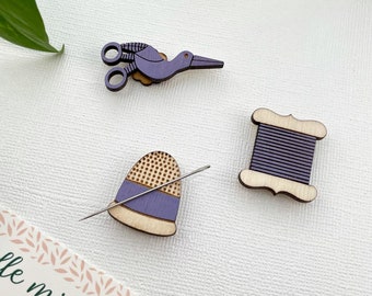May include: Purple wooden needle minder set with a bird, a thimble, and a spool of thread. The set is on a white background with the words "needle minder" written on a piece of paper.