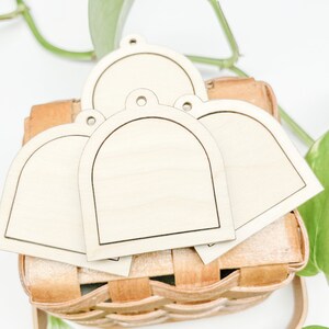 Könnte beinhalten: Vier unfertige Holz-Tags in Bogenform mit einem Loch oben, gestapelt in einem Weidenkorb. Die Tags sind leer und bereit zum Dekorieren.