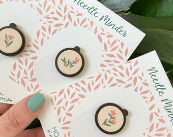 May include: Three wooden needle minders with embroidered floral designs. Each minder is circular and has a brown wooden frame with a white fabric center. The embroidery features a single pink flower with green leaves. The minders are displayed on white cards with the text "Needle Minder" and "HOOPS n' more".