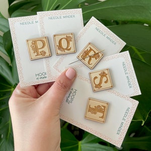 Peut inclure: Cinq protège-aiguilles en bois avec des motifs floraux et les lettres P, Q, R, S et T. Chaque protège-aiguille est sur une carte avec le texte "Needle Minder" et "HOOI n' more".