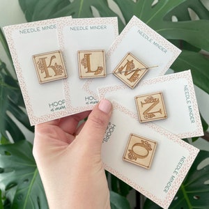 May include: Five wooden needle minders with floral designs and letters K, L, M, N, and O. Each minder is on a white card with the text "NEEDLE MINDER" and "HOOP n' more".