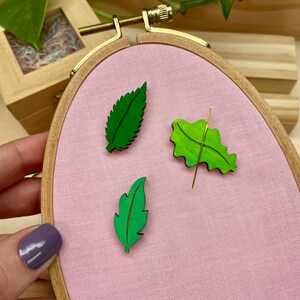May include: Three green wooden leaves with a painted finish are displayed on a pink fabric background inside a wooden embroidery hoop.