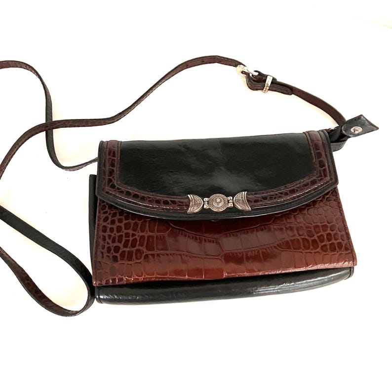 May include: A crossbody bag with a black top flap and a brown, crocodile-textured body. The bag features a decorative silver-toned clasp and a brown adjustable strap. The bag is set against a white background.