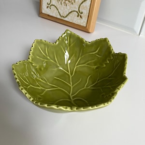 May include: A glossy olive green ceramic bowl shaped like a large maple leaf. The bowl features a detailed embossed vein pattern across its interior surface, with a scalloped, serrated edge that mimics the natural contours of a botanical leaf.