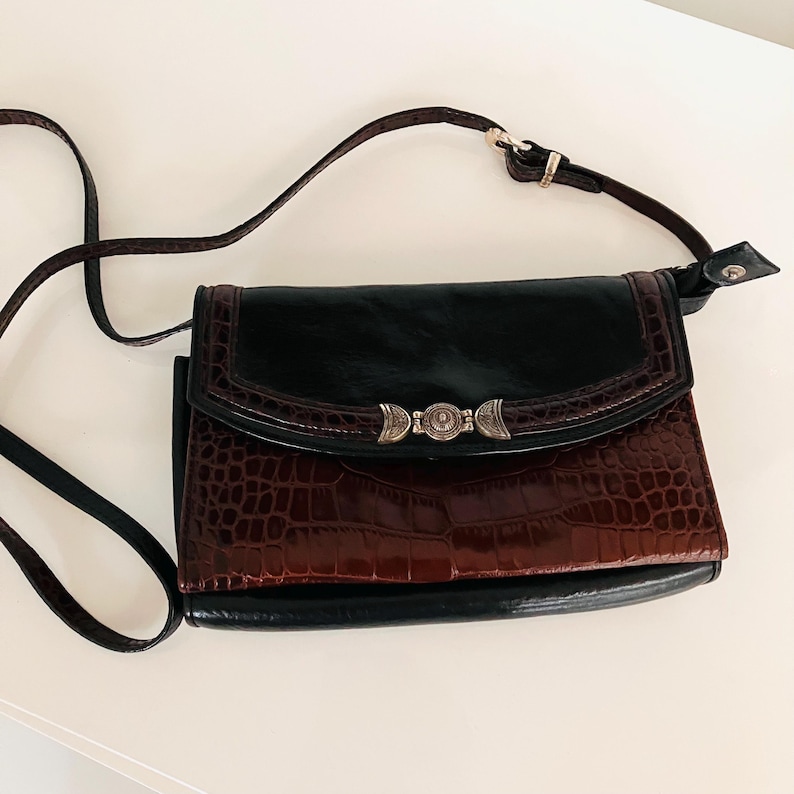 May include: A black and brown crossbody bag with a crocodile-effect texture on the lower section. It has a flap closure with a decorative silver clasp and a long, adjustable strap. The bag is suitable for everyday use.