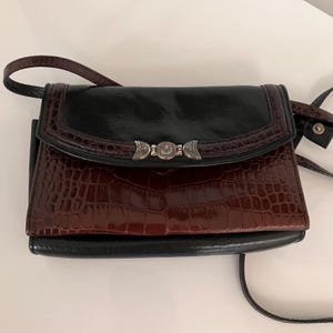 May include: A black and brown crossbody bag with a silver clasp. The bag has a glossy black top flap and a textured, reddish-brown front. The strap is black and the bag is rectangular.