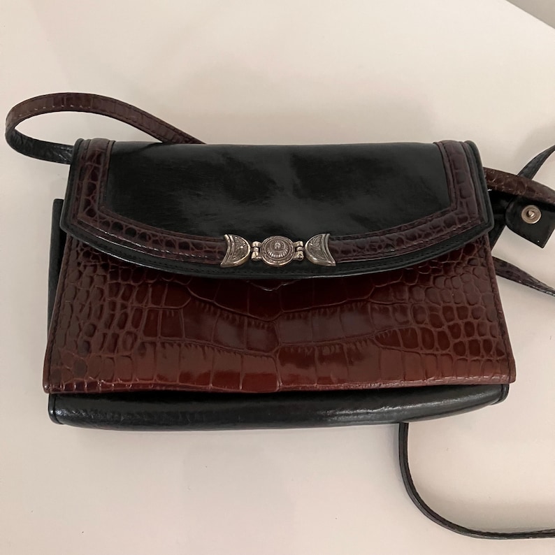 May include: A black and brown crossbody bag with a silver clasp. The bag has a glossy black top flap and a textured, reddish-brown front. The strap is black and the bag is rectangular.