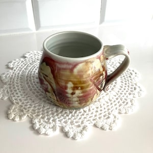 May include: A ceramic mug with a rounded body and a handle. The mug has a white glaze with a pink and brown marbled design.