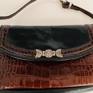 May include: A brown and black leather handbag with a crocodile pattern. The bag has a silver clasp and a black flap. The strap is brown leather. The bag is a clutch style.
