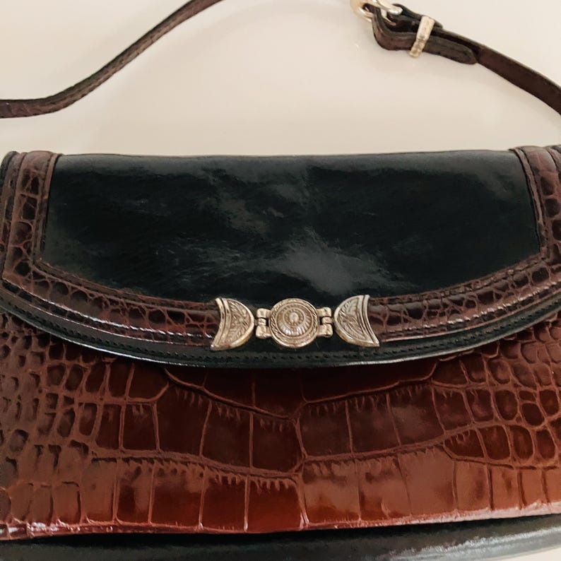 May include: A brown and black leather handbag with a crocodile pattern. The bag has a silver clasp and a black flap. The strap is brown leather. The bag is a clutch style.