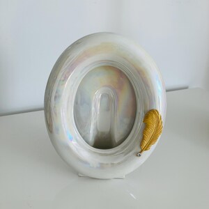 Peut inclure: Un cadre photo de forme ovale avec une finition blanche irisée et brillante qui reflète plusieurs couleurs. Une décoration en métal doré en forme de plume texturée est fixée sur le côté inférieur droit, ornée d'un petit cristal transparent.