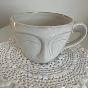 Peut inclure: Une tasse en céramique blanc cassé brillant présentant un visage humain sculpté sur l'extérieur. La tasse a une forme de bol large et arrondie, une finition lisse et une anse incurvée sur le côté droit, avec des traits du visage en relief.