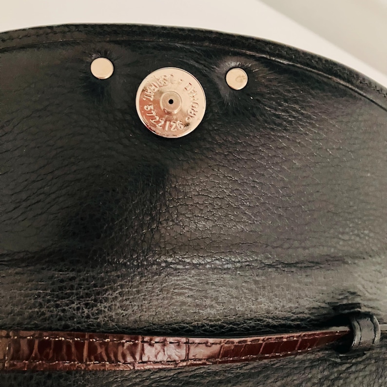 May include: Close-up of a black leather handbag flap with a magnetic closure. The flap features a silver magnetic snap and two silver rivets. A dark brown, textured leather strap is visible at the bottom.