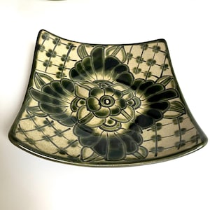 May include: A square ceramic dish with curved sides featuring a dark green floral pattern centered on a light beige background. The design includes a grid of thin dark lines with small dots at the intersections, framing the central flower motif.