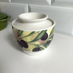 Peut inclure: Un beurrier en céramique avec un couvercle et une base blancs, orné d'un motif décoratif représentant des olives violet foncé et des feuilles vertes sur un fond jaune pâle et crème. Le récipient présente une forme arrondie et bombée.