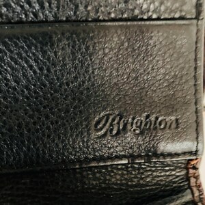 May include: A close-up shot of a black leather wallet with a textured finish. The word "Brighton" is embossed on the front. The wallet appears to be new.