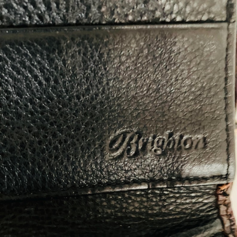 May include: A close-up shot of a black leather wallet with a textured finish. The word "Brighton" is embossed on the front. The wallet appears to be new.