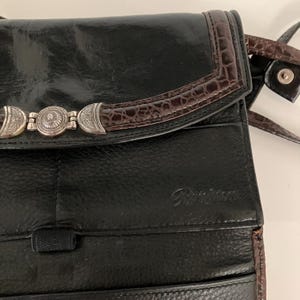 May include: Black leather crossbody bag with a brown faux-crocodile trim and silver decorative accents. The bag has a long strap and the brand name "Brighton" embossed on the front.