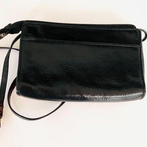 May include: A black crossbody bag with a glossy finish. The bag is rectangular with a zipped top and a front pocket. It has a long, thin strap for shoulder or crossbody wear.