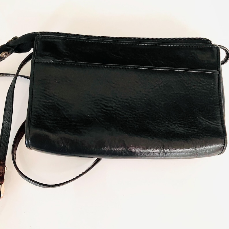 May include: A black crossbody bag with a glossy finish. The bag is rectangular with a zipped top and a front pocket. It has a long, thin strap for shoulder or crossbody wear.
